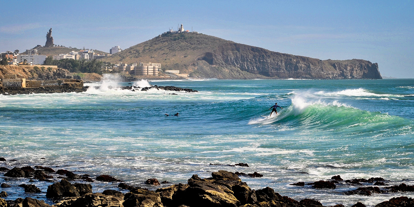 Un surfer prend une vague à Dakar, avec la colline des Mamelles