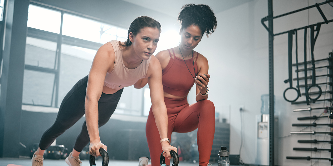 Personal trainer, kettle bell and stopwatch with a black woman coaching a client in a gym for her workout. Health, exercise or training and a female athlete doing a plank with her coach taking time